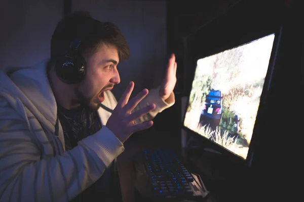 Jogador aterrorizado joga jogos no seu computador. Um jovem emocional nos fones de ouvido joga jogos em um computador. Conceito de jogador . — Fotografia de Stock