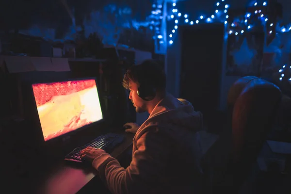 A focused gamer sits at the computer at home in the cozy room an — Stock Photo, Image