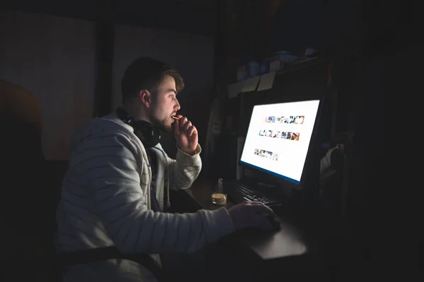 Мужчина сидит дома за компьютером ночью и ест печенье. Перекусить за компьютером — стоковое фото