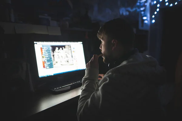 Un hombre se sienta de noche en un escritorio de computadora usando Internet. Navegación nocturna en Internet. Gamer se sienta de noche en una mesa cerca de un ordenador con galletas en las manos y mira el monitor — Foto de Stock