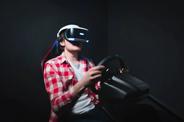 Hráč hraje Vr závod na černém pozadí. Virtuální realita na pronájem simulátoru. Člověk sedí za auto simulátor a hraje hry v Vr brýle. — Stock fotografie