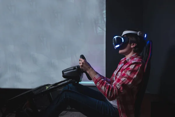 Gamer joue VR course sur un fond noir. Réalité virtuelle sur simulateur de voiture. Un homme est assis derrière un simulateur de voiture et joue à des jeux en lunettes VR . — Photo