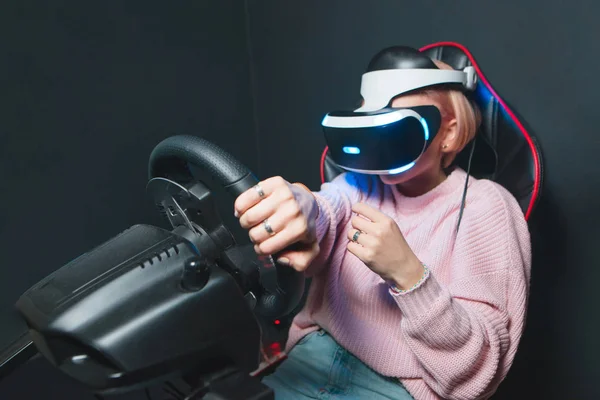 La fille est assise dans le simulateur d'une voiture avec un casque BP sur la tête et joue le jeu. La femme joue la course dans une réalité virtuelle sur un fond noir . — Photo