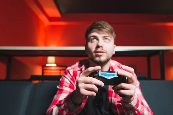 Homem com barba sentado em uma sala com um gamepad em suas mãos e jogando jogos. Um jogador em uma camisa colorida brilhante joga o jogo no console . — Fotografia de Stock