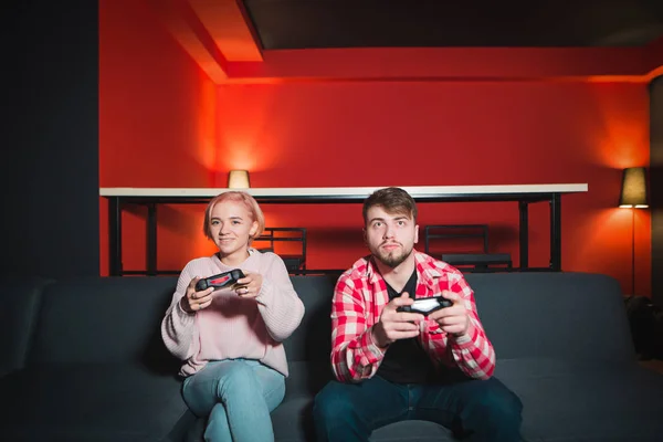 Couple of young people, a man and a girl playing video games on the console. A couple is sitting on the couch with joysticks in her hands and playing games
