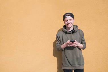 Elleri ve gülümser bir telefon ile turuncu bir duvar arka plan üzerinde gülümseyen genç bir adam duruyor. Kamera bir göz.
