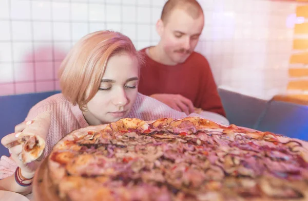La chica huele el aroma de la pizza fresca y sabrosa. Pareja joven sentada en una pizza cerca de una pizza grande. Deliciosa pizza y gente —  Fotos de Stock