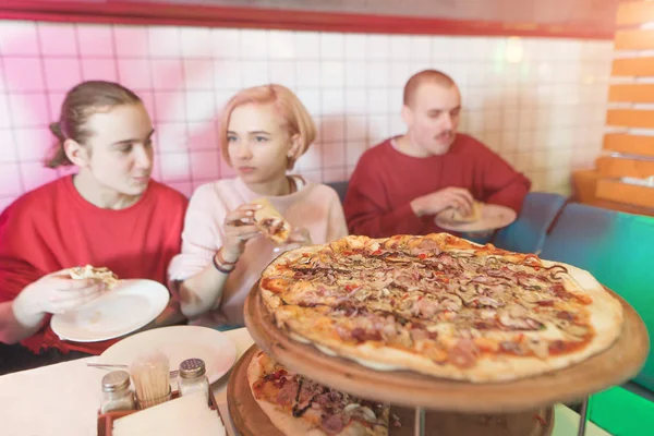 Lezzetli büyük bir pizza insanlar arka plan üzerinde. Bir grup genç insan rahat pizzacıda oturur ve lezzetli pizza yiyor — Stok fotoğraf