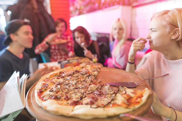 Gran pizza de aperitivo en el fondo de un grupo de amigos. Un grupo de jóvenes estudiantes sentados en una pizzería y comiendo pizza. Enfoque en pizza — Foto de Stock