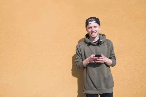 Um jovem sorridente está no fundo de uma parede laranja com um telefone nas mãos e sorri. Um olhar para a câmera . — Fotografia de Stock