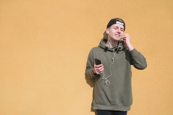 Портрет молодого человека на фоне оранжевой стены, слушающего музыку в наушниках. Подросток в кепке стоит на фоне цветной стены и слушает музыку . — стоковое фото