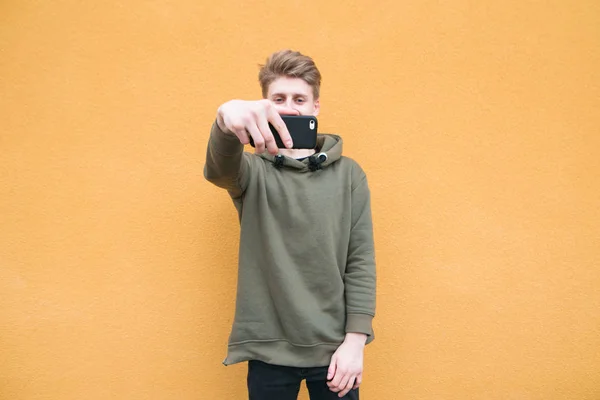 Gelukkig student in casual kleding neemt selfie op de achtergrond van een oranje muur. Een jonge man neemt een foto op een smartphone — Stockfoto