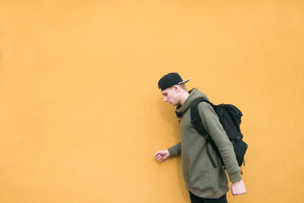 Um jovem vestido com roupas casuais e uma mochila nas costas está no fundo de uma parede laranja — Fotografia de Stock