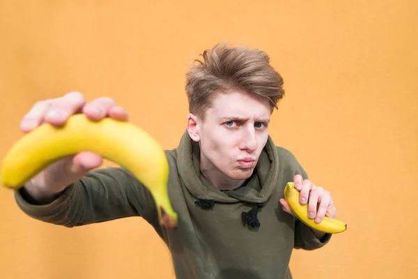 A cute and funny young man with bananas in his hands posing on an orange background with bananas in his hands. A guy with a banana looks at the camera