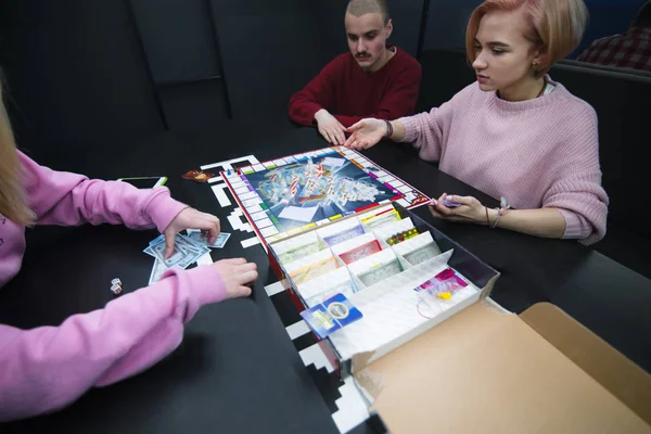 Los jóvenes se sientan a la mesa y juegan un monopolio. Juegos de mesa con amigos . — Foto de Stock