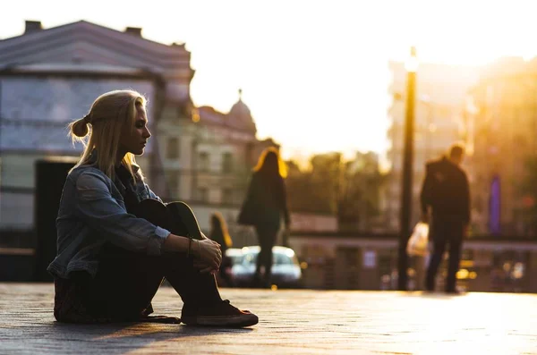 A menina loira se senta no fundo da cidade e do pôr do sol . — Fotografia de Stock