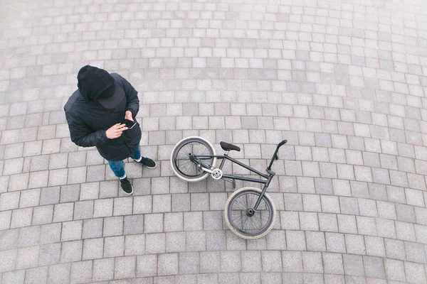 A young man stands on the street near a BMX bike and uses a smartphone. Rest after tricks on BMX. Top view