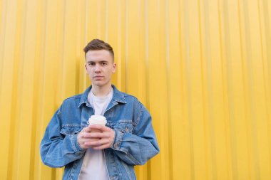 Genç bir adam sarı bir duvar arka planı elinde bir fincan kahve ile standları ve kameraya benziyor