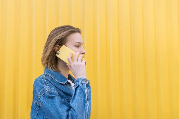Ένα κομψό κορίτσι μιλά για ένα κίτρινο τηλέφωνο στο φόντο των έναν κίτρινο τοίχο και κοιτάζει προς την πλευρά. Επικοινωνία μέσω τηλεφώνου — Φωτογραφία Αρχείου