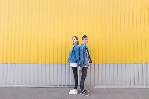 Retrato de um jovem casal em altura máxima contra um fundo de uma parede amarela. Um par de hipsters em jaquetas de ganga posa contra o fundo da parede — Fotografia de Stock