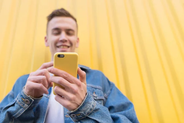 Ein fröhlicher junger Mann genießt sein Smartphone in einer hellen Hülle auf gelbem Hintergrund. Ein lächelnder junger Mensch benutzt ein Smartphone auf dem Hintergrund einer gelben Wand. Fokus auf das Smartphone — Stockfoto