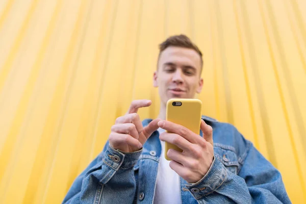 Feliz joven vestido con chaqueta de mezclilla se comunica con amigos en Internet con un teléfono inteligente en el fondo de una pared amarilla. Concéntrate en el smartphone —  Fotos de Stock