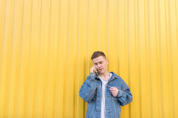 Een knappe jongeman praten op een smartphone op de achtergrond van een gele muur. Hipster in een jeans jasje roept de smartphone op een gele achtergrond — Stockfoto