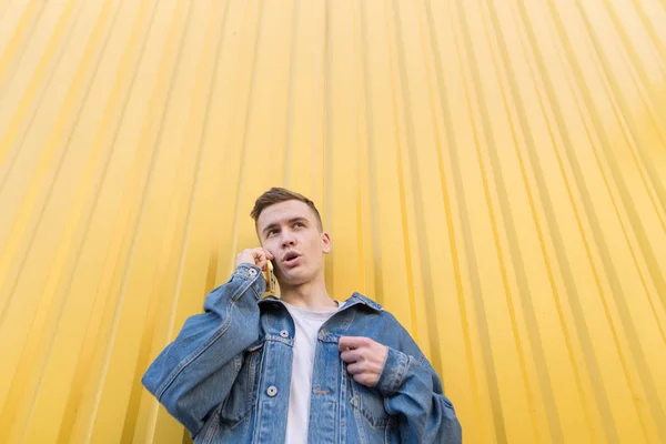Hipster speaks by telephone on the background of a yellow wall. Portrait of a man in a denim jacket while talking on the phone — Stock Photo, Image