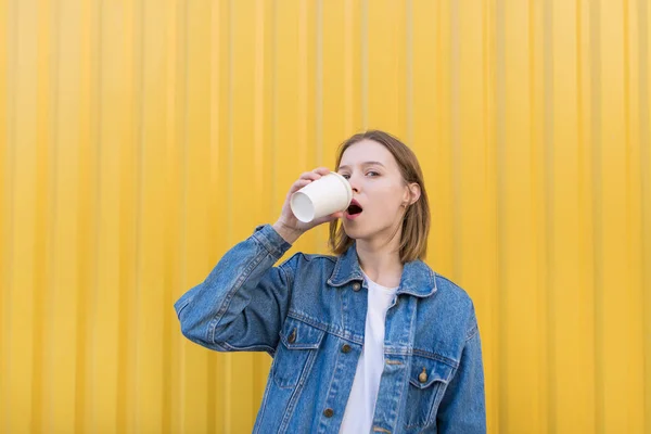 Uma linda garota fica no fundo de uma parede amarela e bebe café de uma xícara. Uma jovem em uma jaqueta de ganga bebe café de uma xícara em um fundo amarelo — Fotografia de Stock