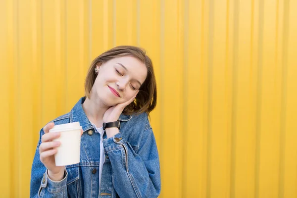 Jolie fille avec une tasse de café se tient sur un fond de mur jaune. Belle fille sur un fond jaune cette tasse de café dans ses mains . — Photo