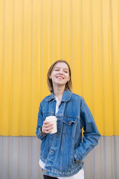 Menina feliz está de pé sobre um fundo amarelo e beber café. Retrato de uma menina com os olhos fechados e com uma xícara de café nas mãos atrás do fundo da parede amarela . — Fotografia de Stock