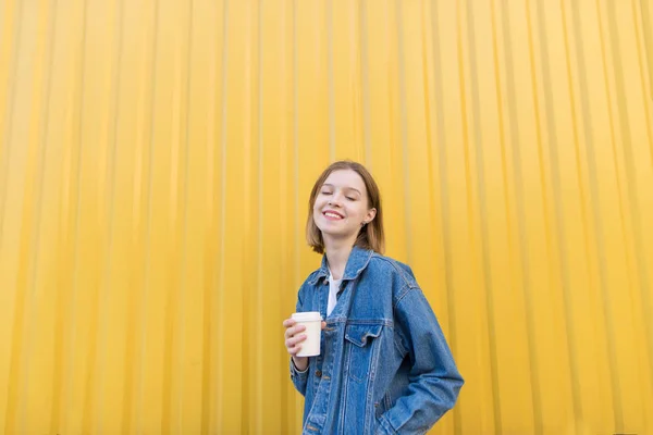 Menina feliz com uma xícara de café em um fundo amarelo. Retrato de uma menina com os olhos fechados e com uma xícara de café nas mãos atrás do fundo da parede amarela . — Fotografia de Stock