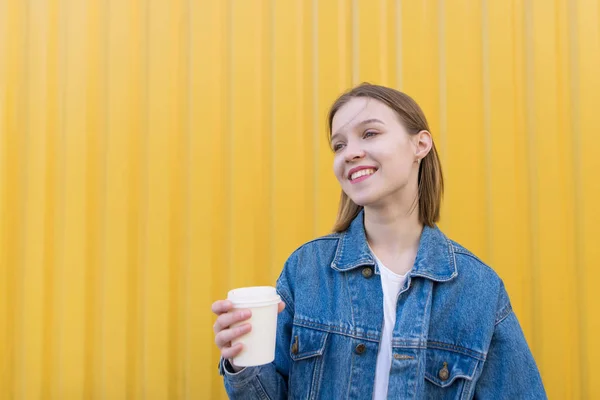 Uma garota atraente com uma xícara de café em suas mãos fica no fundo de uma parede amarela e sorri. Menina feliz fica em um fundo amarelo e olha para o lado — Fotografia de Stock