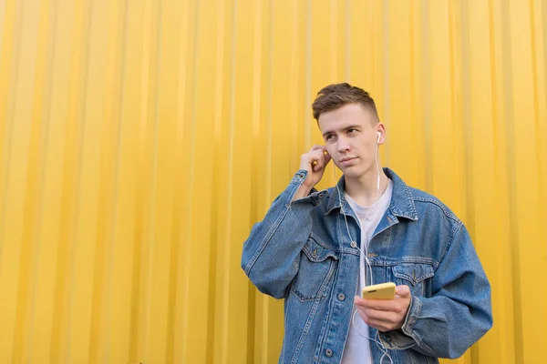 Um homem bonito ouve música nos fones de ouvido e segura o telefone no fundo da parede amarela. Olha para o lado. Espaço de cópia . — Fotografia de Stock