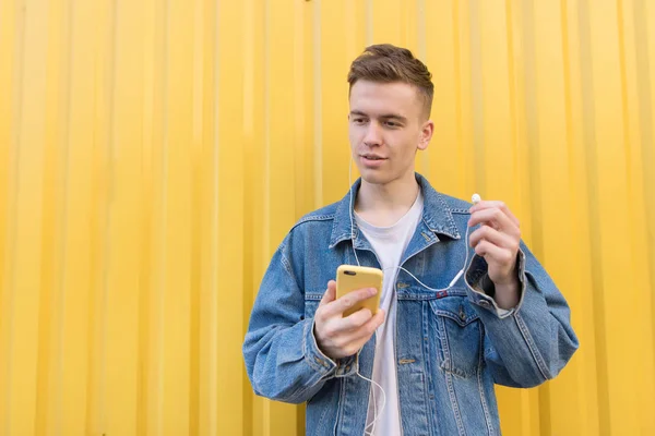 Молодий чоловік слухає музику на жовтому тлі. Щасливий студент стоїть зі смартфоном в руках на тлі жовтої стіни і вставляє навушники в його вуха — стокове фото