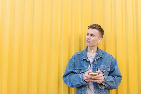 Молодой человек в джинсовой куртке слушает музыку в наушниках, держит телефон в руках и смотрит в сторону. На фоне желтой стены. Copyspace . — стоковое фото