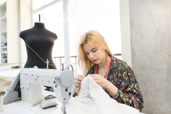 Una ragazza sarta siede sul posto di lavoro vicino a una macchina da cucire e cuce un panno bianco con le mani. Fare vestiti al tuo posto di lavoro. Concetto di cucito — Foto Stock