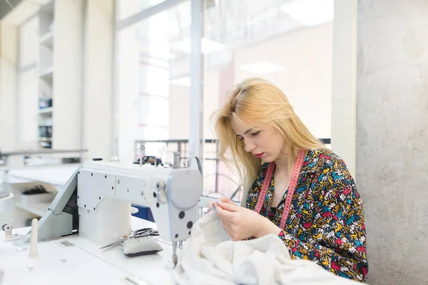 Una costurera linda trabaja en el lugar de trabajo cerca de una máquina de coser. Lugar de trabajo para costureras. La chica está cosiendo su ropa en casa —  Fotos de Stock