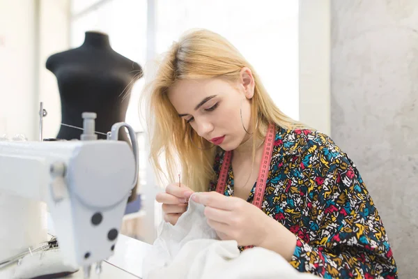 El retrato de una costurera se sienta en el lugar de trabajo cerca de una máquina de coser y cose un paño con las manos. Trabajo de costureras. Sastre del lugar de trabajo - máquina de coser, maniquí, tela, aguja . — Foto de Stock