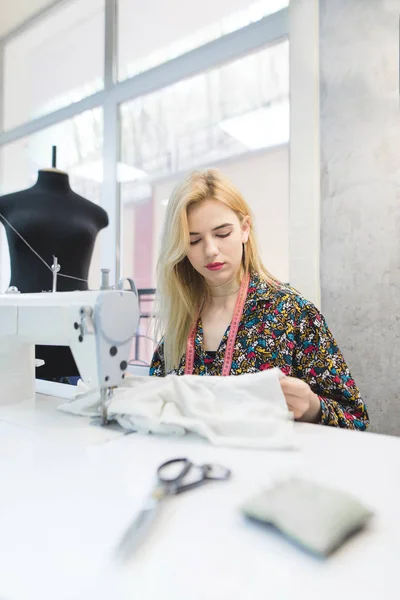 Портрет молодой швеи, работающей с тканью на рабочем месте возле швейной машинки. Молодая портниха шьет одежду на швейной машинке — стоковое фото