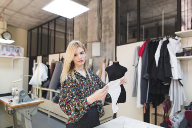 Güzel bir terzi onun elinde beyaz bir bez ile bir stüdyoda duruyor ve kameraya benziyor. Moda giysiler oluşturulması
