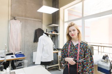 Genç güzel terzi makas elinde işyeri ve kamera için poz arka planı var. Giysi yaratma çalışma süreci. Güzel bir terzi portresi