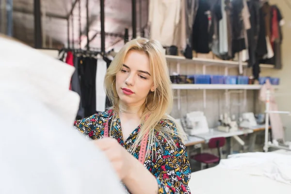 Retrato de un joven diseñador de moda en el trabajo. Joven modista mujer se encuentra en un estudio cerca de una manicura con ropa y obras. Crear ropa elegante — Foto de Stock