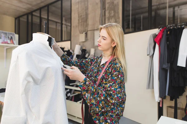 Un joven diseñador de moda crea ropa en un estudio de moda. Mujer joven modista se encuentra en un estudio bien cuidado y ajusta la ropa. Concepto de negocio de costura . — Foto de Stock