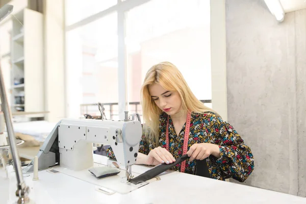 Una costurera linda se sienta en el lugar de trabajo y trabaja en una máquina de coser en un estudio brillante. Lugar de trabajo del sastre . — Foto de Stock