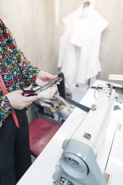 Costurera de pie en el lugar de trabajo y corta la tela con tijeras. Sastre del lugar de trabajo - máquina de coser, maniquí, tela, aguja . —  Fotos de Stock