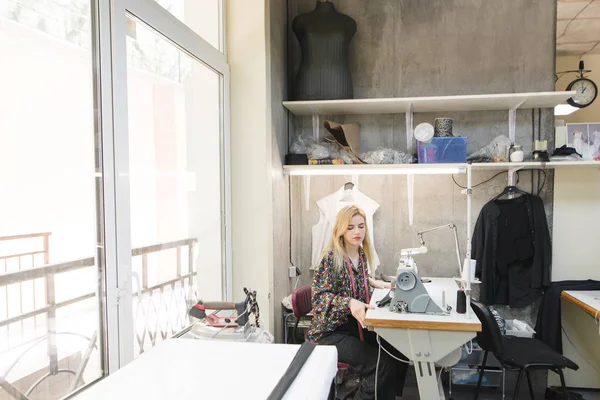 Estudio de ropa de moda. Crear ropa elegante. Una costurera linda en el lugar de trabajo en un estudio moderno y brillante. Trabajo de modista en la máquina de coser. Concepto de negocio de costura . —  Fotos de Stock