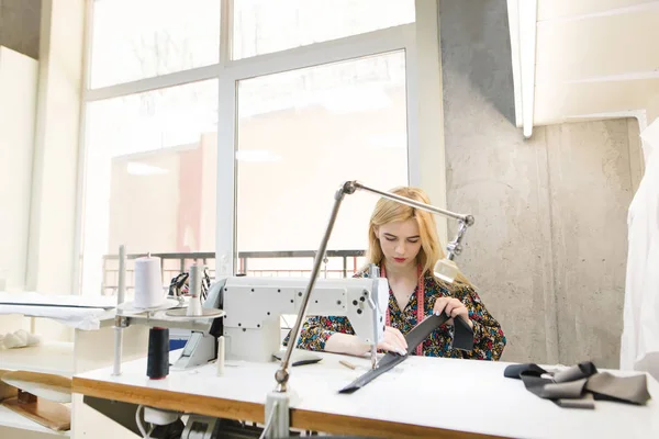 Retrato de una linda costurera joven en el trabajo en una máquina de coser profesional en un salón de luz diseñando ropa. Concepto de costurera. Creación de ropa de diseño en un estudio moderno y luminoso . — Foto de Stock