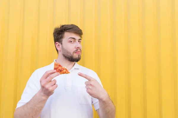 A beautiful bearded man stands with a piece of pizza on the background of a yellow wall and looks at the camera. The man shows his finger on a piece of pizza