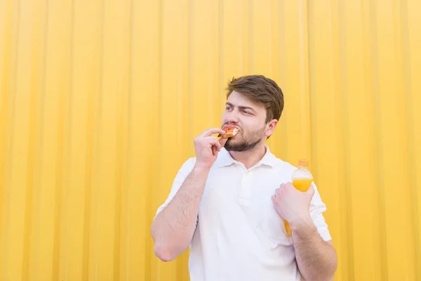 Un buen hombre come sobre un fondo de pared amarilla. Un estudiante come un pedazo de pizza sobre un fondo amarillo y sostiene una botella de bebida en sus manos . — Foto de Stock